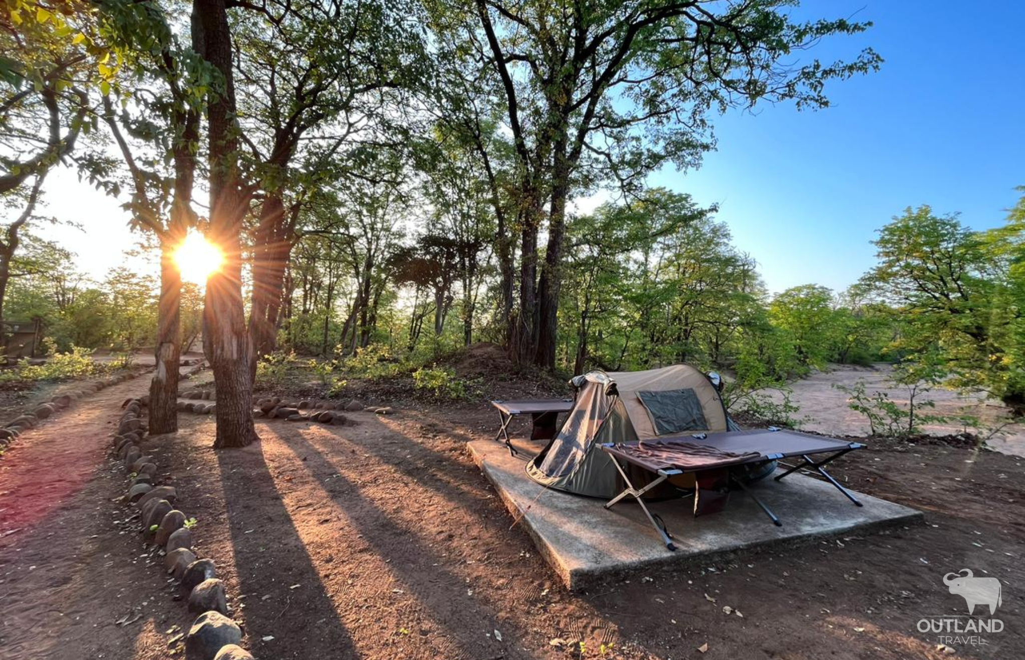 Wilderness camping in a tent with stretchers in Botswana with the sun setting behind beautiful trees - Outland Tavel