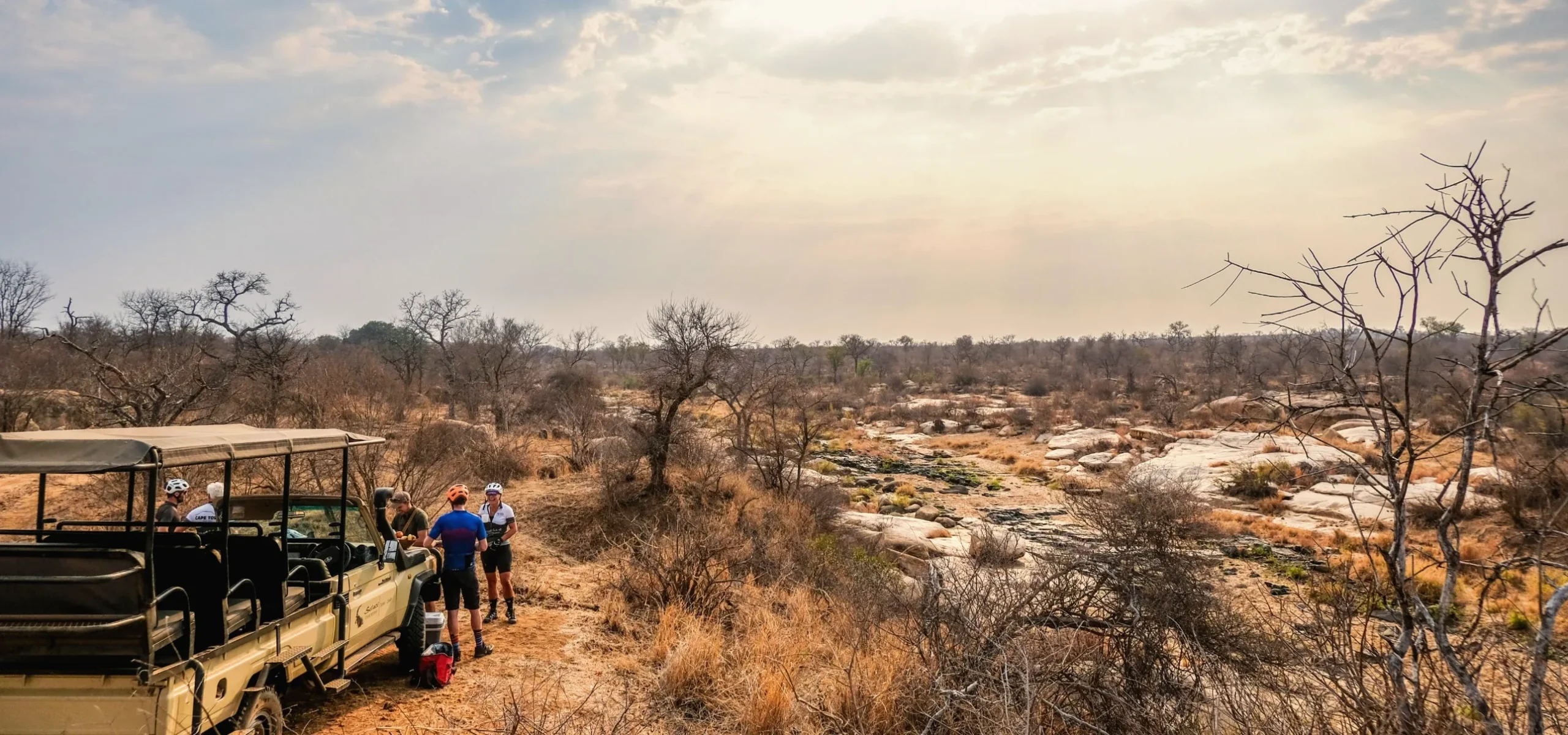 Cyclists enjoying a sunrise bush coffee stop beside an open safari vehicle overlooking the rocky Selati River in Big 5 country.
