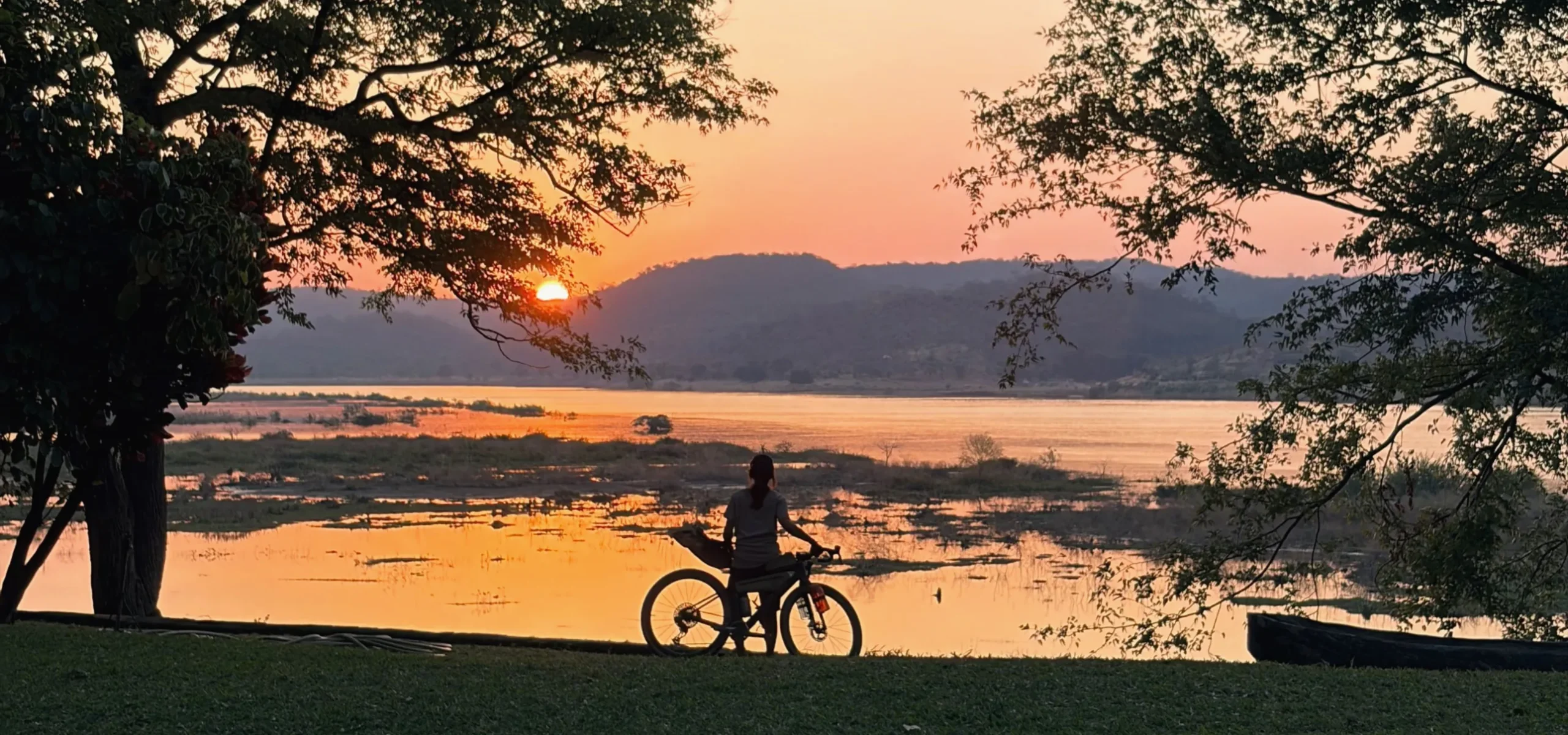 Silhouette of a cyclist standing with her bike on the riverbanks of the Zambezi as the sun sets behind the hills.
