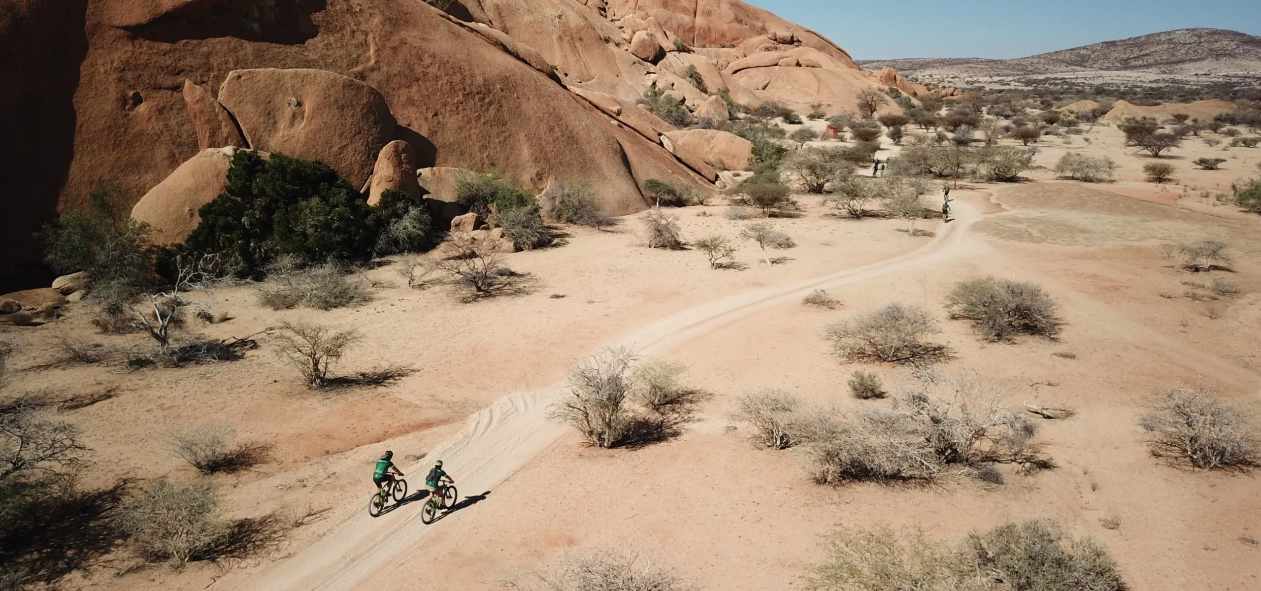 Aerial view of mountain bikers riding a sandy track past towering red granite domes in the Namibian desert.